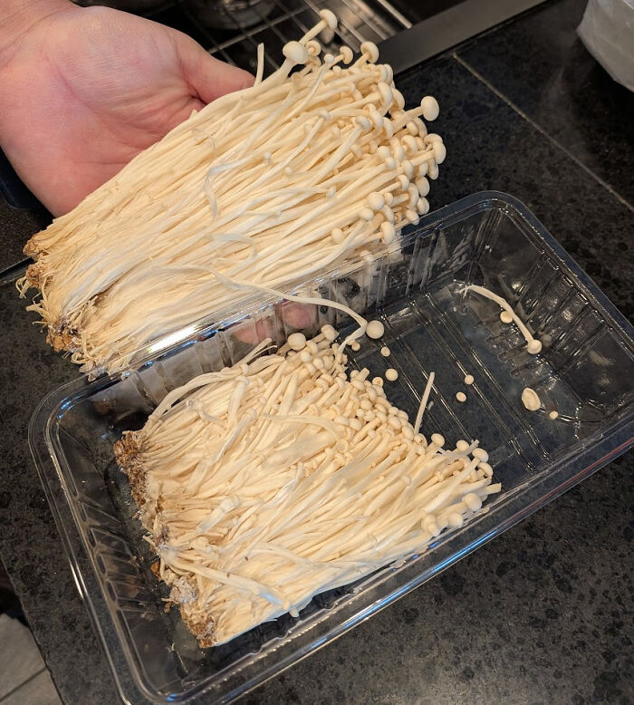 Hand holding a smaller portion of enoki mushrooms labeled with shrinkflation showing less product in clear plastic packaging.