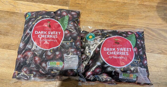 Two bags of frozen dark sweet cherries by Sainsbury's on a wooden surface showing shrinkflation difference in size.