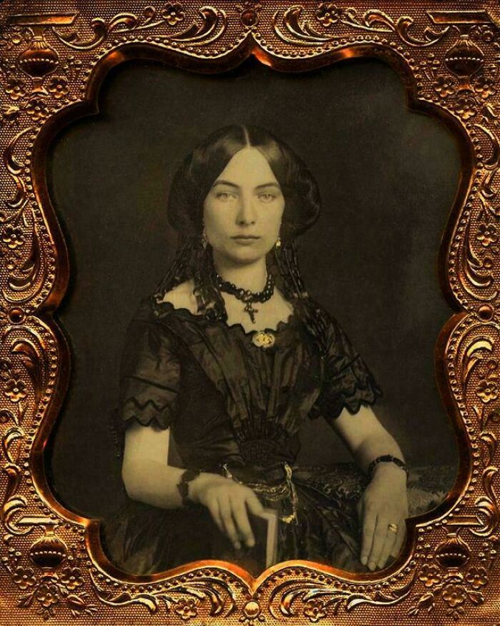 Victorian-era woman in an ornate historical garment, seated with jewelry, holding a book in a decorative vintage frame.