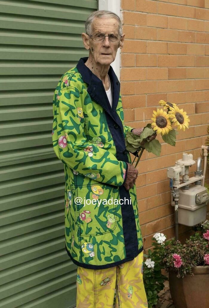 Elderly man wearing a vibrant green patterned historical garment holding sunflowers against a brick and green wall background.
