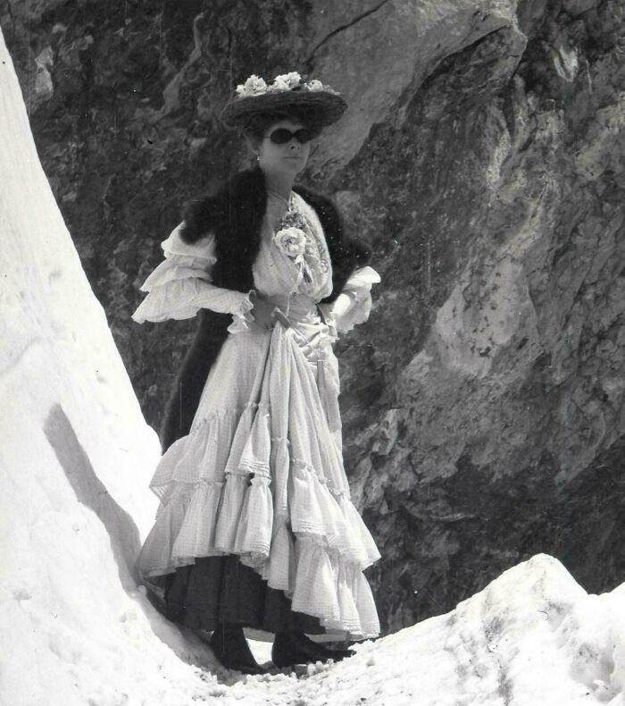 Woman wearing historical garments with layered dress and fur shawl, standing on snowy rocky terrain in vintage style.