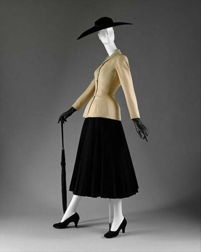 Vintage historical garment with beige fitted jacket, black pleated skirt, wide-brim hat, gloves, and umbrella on mannequin.