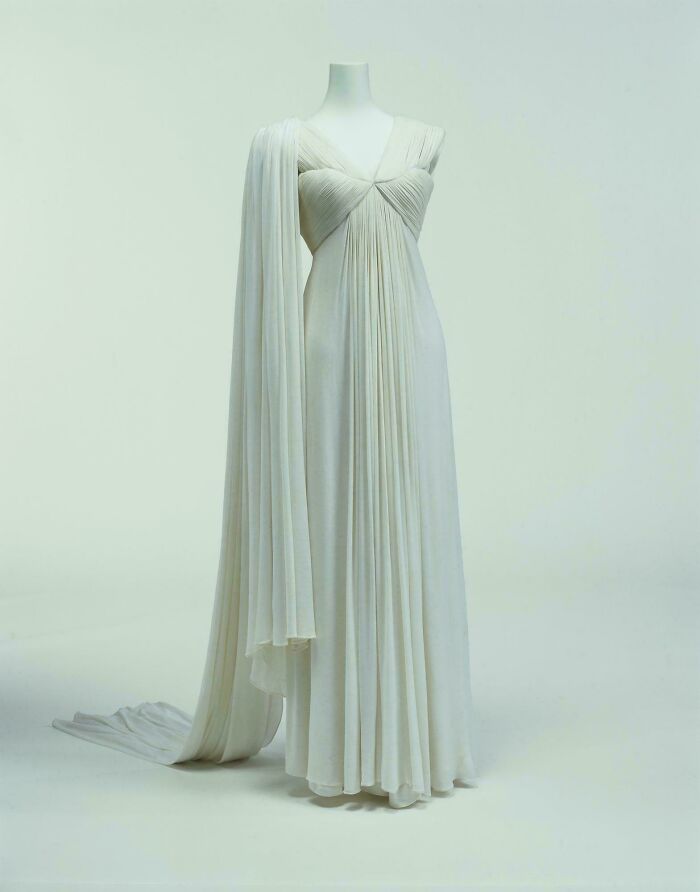 Elegant historical garment in white with flowing pleats and draped fabric displayed on a mannequin.