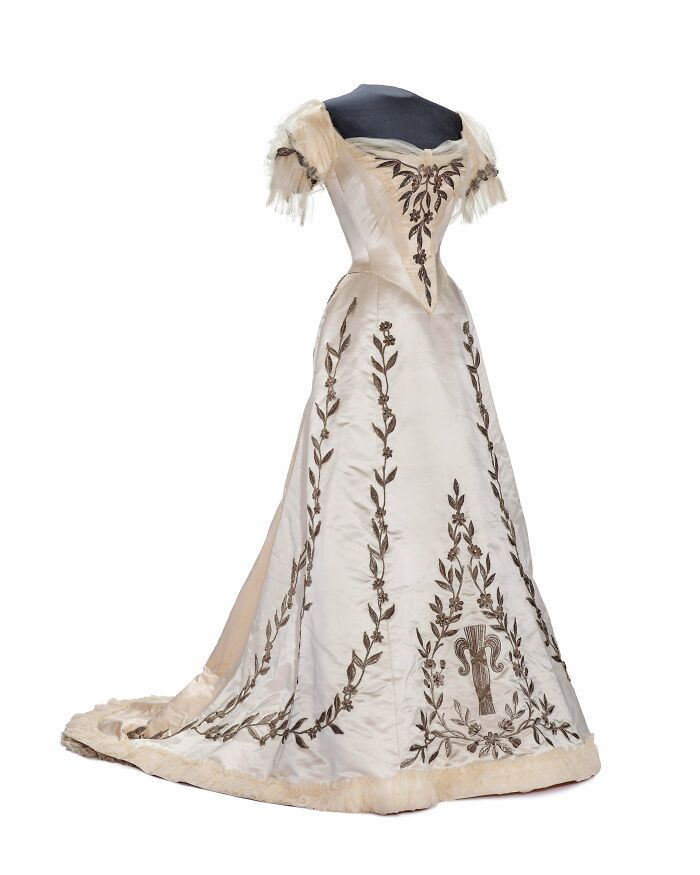 Elegant historical garment with intricate embroidery and a long train displayed on a black mannequin.
