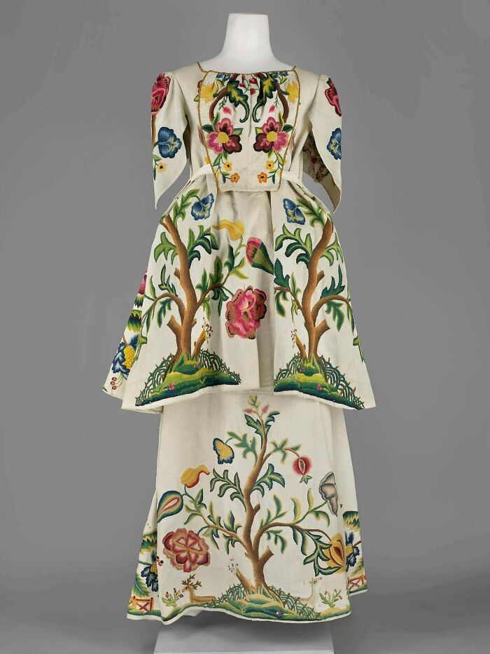 Historical garment with detailed floral embroidery and tree motifs on a cream fabric displayed on a mannequin.