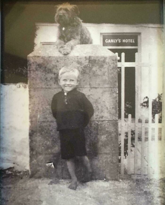 Black and white vintage photo of a smiling boy standing barefoot by a stone wall with a dog lying on top, decades ago.