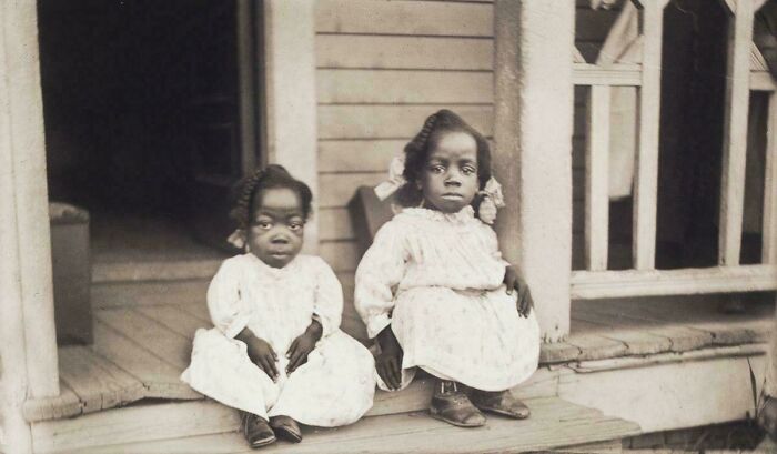 Two young girls in old-fashioned dresses sitting on a wooden porch in vintage black and white photo from decades ago