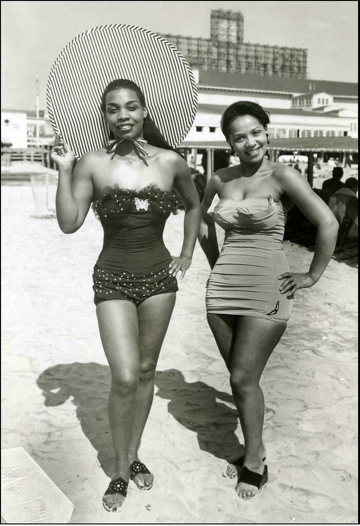 Two women in vintage swimsuits posing on a sunny beach, reflecting decades-old fashion in classic black and white photography.