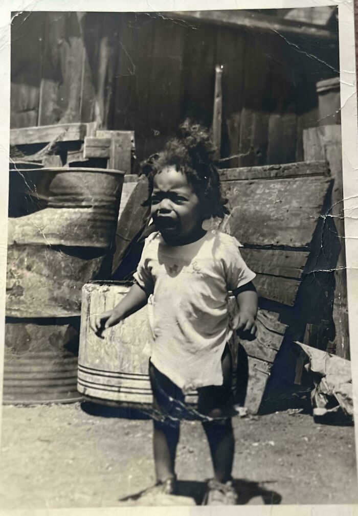 Black and white vintage photo of a young child standing outdoors near old barrels and wooden crates from decades ago.