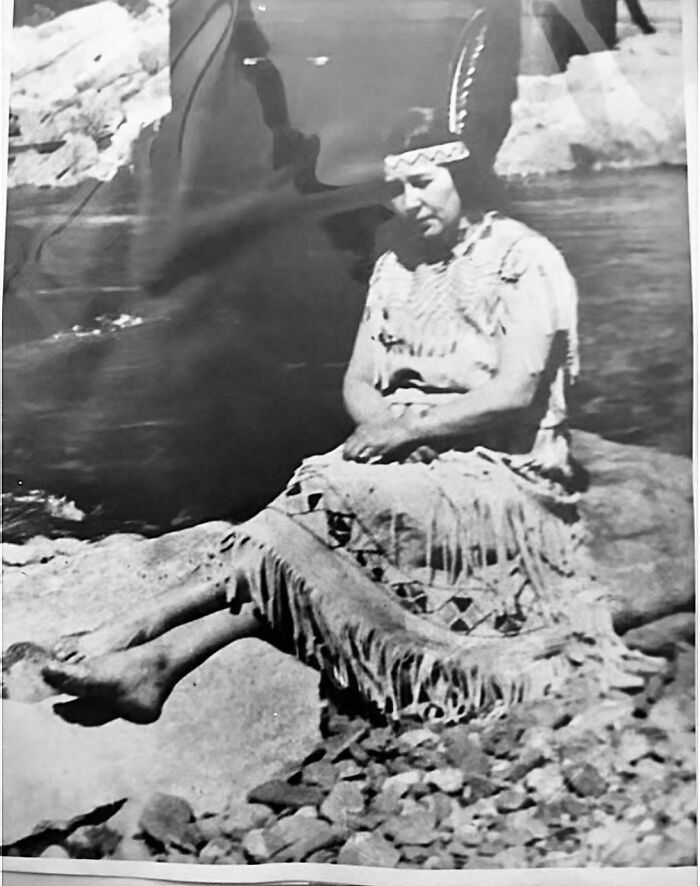 Black and white photo of a Native American woman in traditional clothing sitting on rocks near water vintage photo decades ago