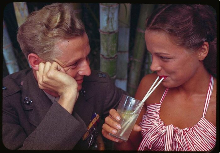 Vintage photo of a man in military uniform gazing at a woman sipping a drink through two straws, capturing moments from decades ago.