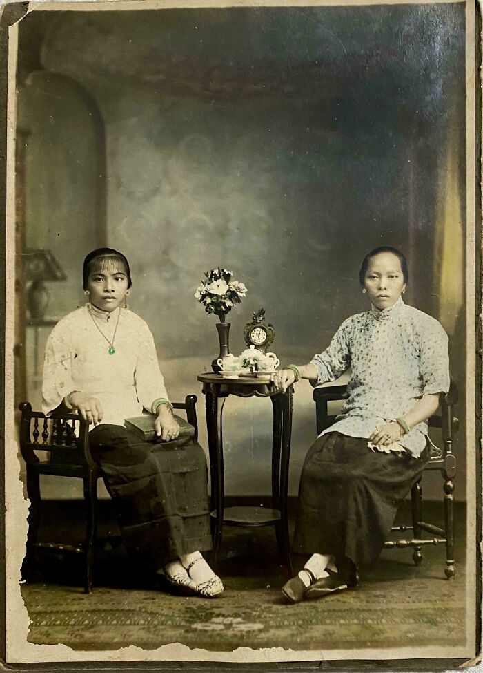 Two women from decades ago seated by a small table with flowers and a clock in a vintage photo from the past.
