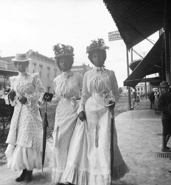 Three women in vintage dresses and hats walking on a street in old black and white photo from decades ago.