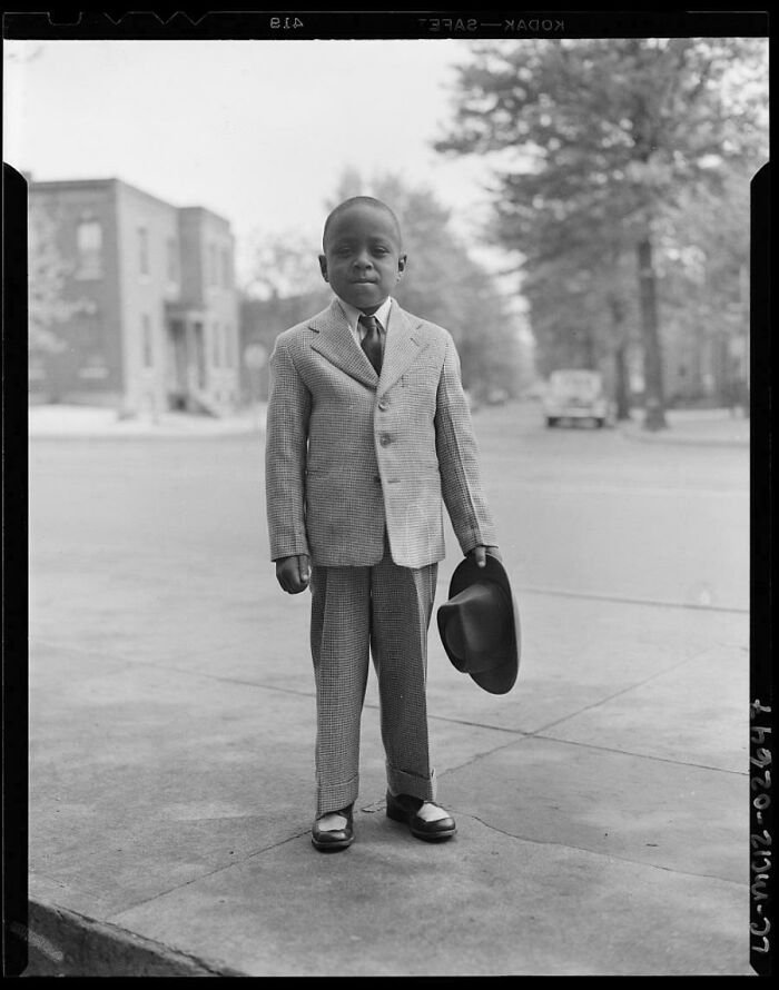 Young boy in a vintage suit holding a hat, standing on a sidewalk in a neighborhood, classic photo from decades ago.