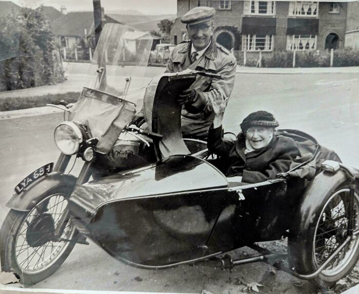 Black and white photo of an elderly couple with a vintage motorcycle and sidecar from decades ago smiling on a suburban street