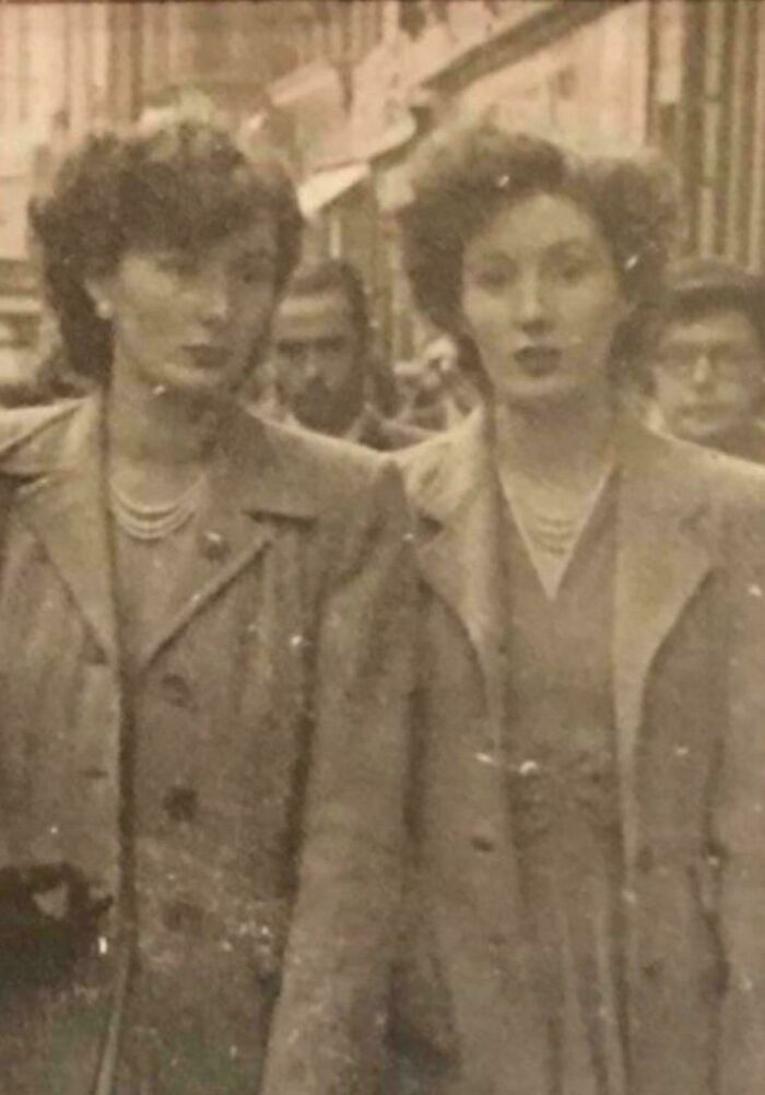 Two women in vintage coats walking on a crowded street in a black and white photo from decades ago.