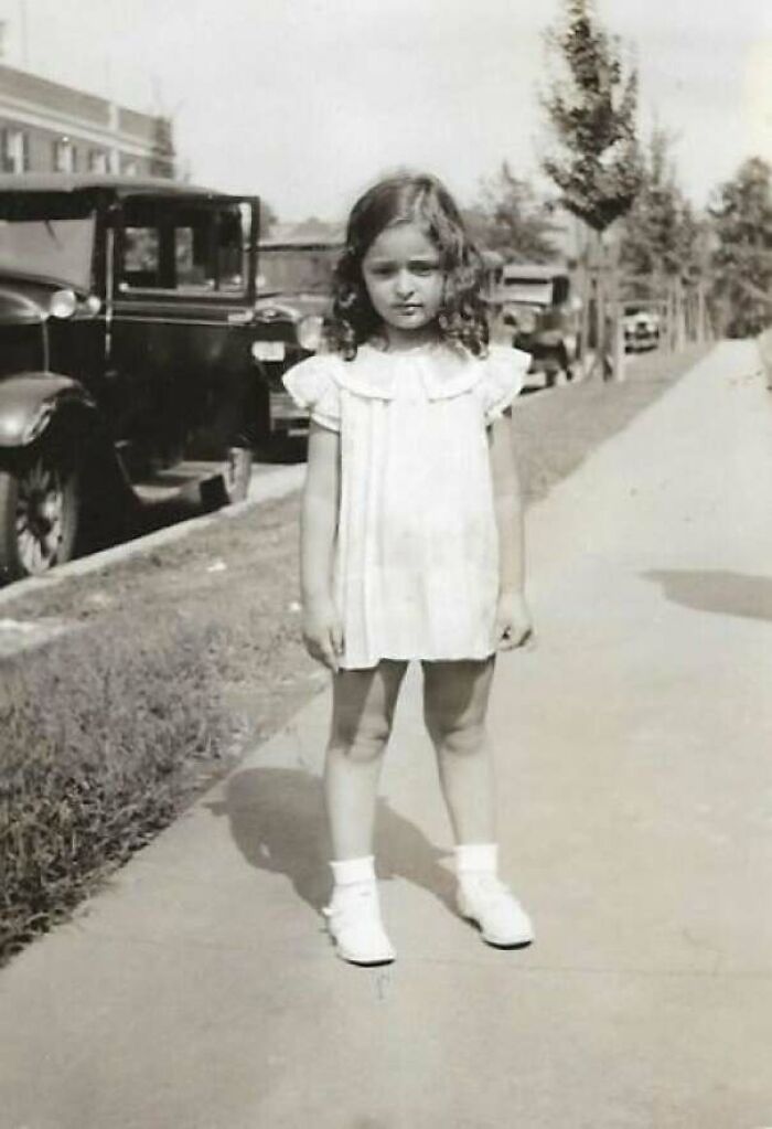 Young girl in vintage dress standing on sidewalk near classic cars in a decades-old photo showing history through images