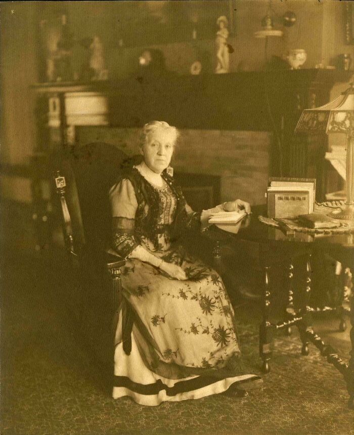 Sepia-toned photo of an elderly woman in vintage dress reading by a table, capturing a moment from decades ago.