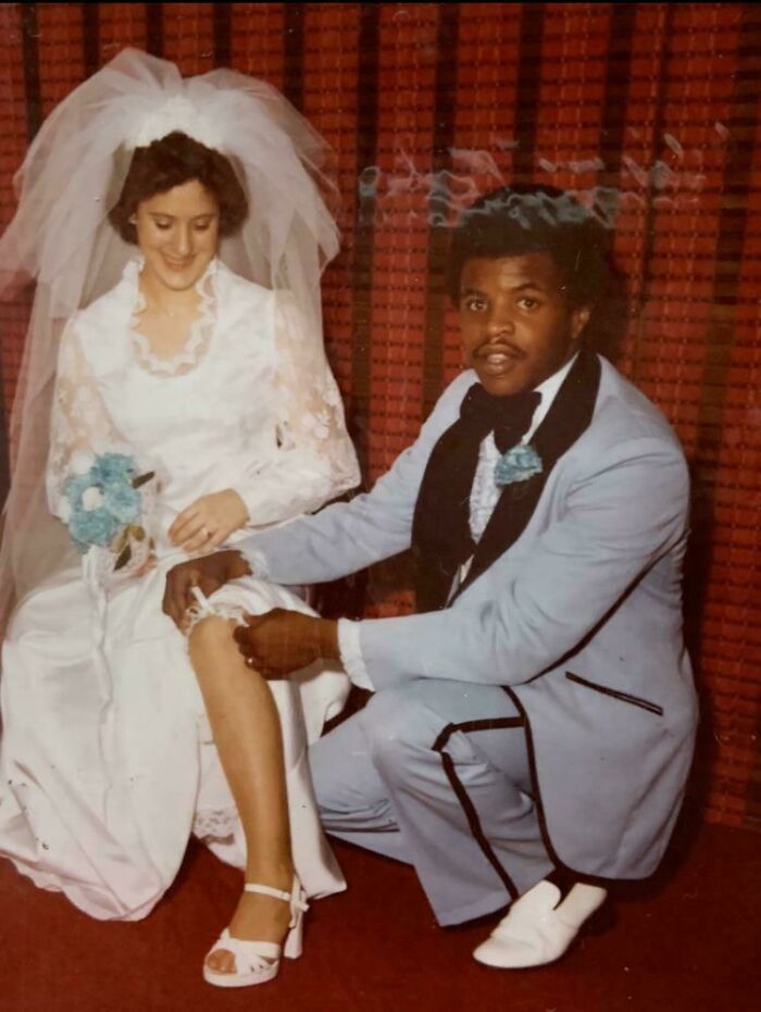 Bride in vintage wedding dress smiling while groom adjusts her garter in a classic photo from decades ago.