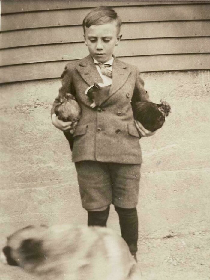 Young boy in vintage clothing holding two chickens in a black and white photo from decades ago.