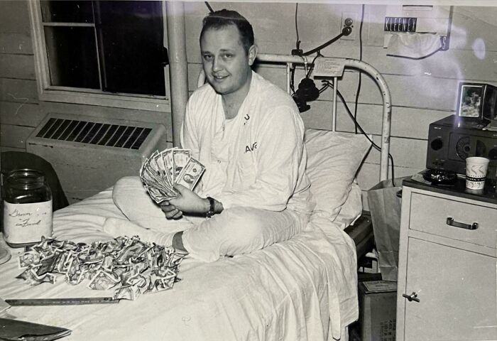 Man sitting on a bed holding vintage cash, surrounded by folded bills from decades ago in a black and white photo.