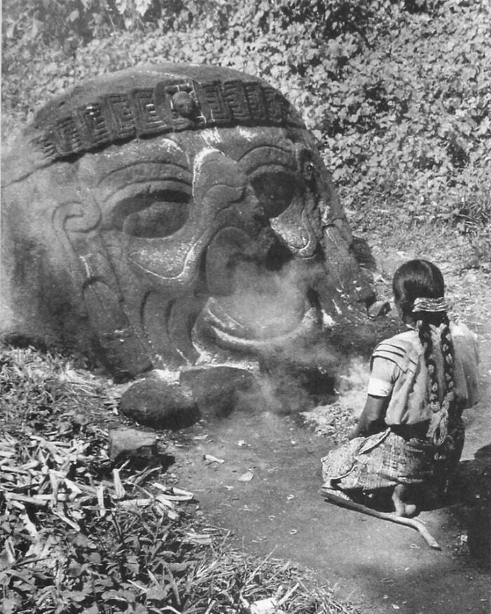 Ancient carved stone artefact with intricate face design, surrounded by vegetation and a person performing a ritual nearby.