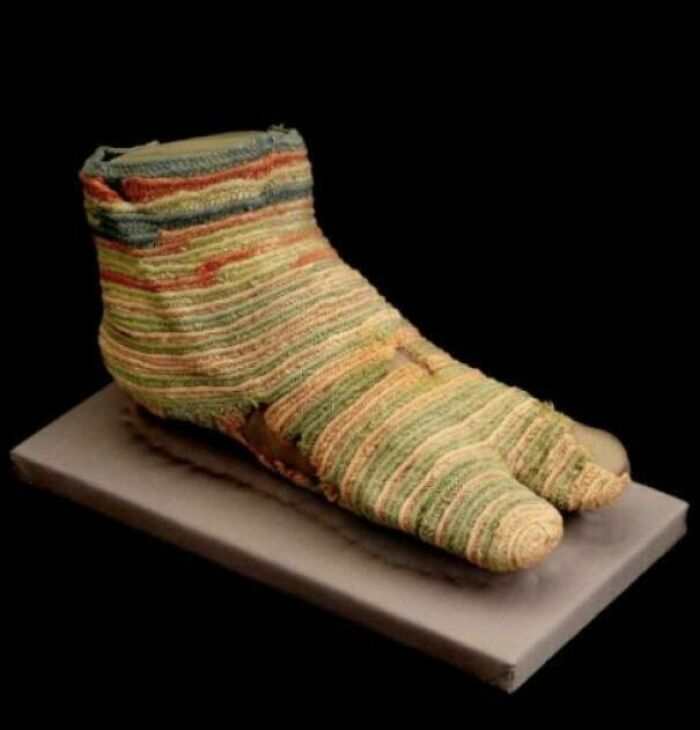 Ancient woven textile shoe artifact with colorful stripes displayed on a platform representing historical craftsmanship.