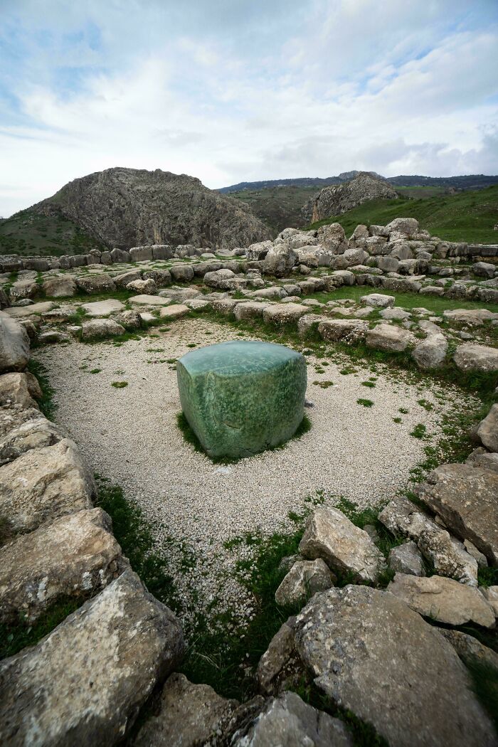 Ancient artefact of a green stone in the center of ruins surrounded by rocks and mountains under a cloudy sky.