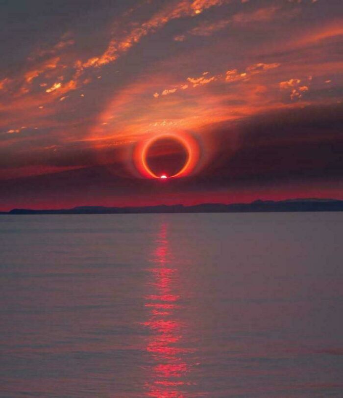 A strange Earth sunset with a glowing red ring around the sun over calm water and a dramatic cloudy sky.