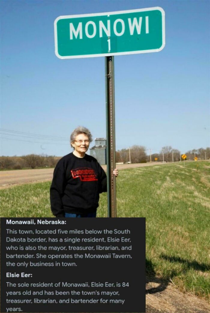 Elderly woman standing beside Monowi town sign, the only resident managing multiple roles in this strange earth location.