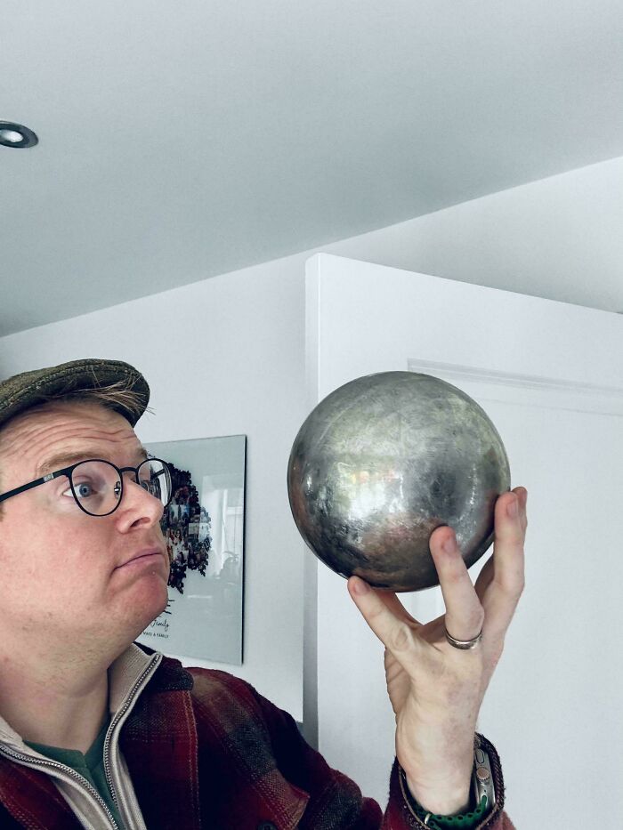 Man wearing glasses and a hat holding a large reflective metal sphere, showcasing strange earth phenomenon indoors.