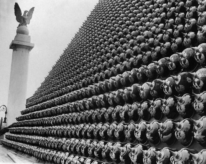 Rows of historical helmets displayed on a large wall next to a tall monument with an eagle statue above it.