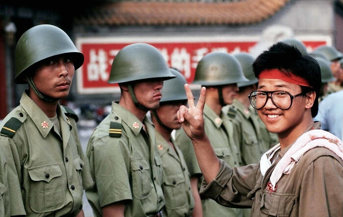 Young man making peace sign in front of soldiers wearing helmets, captured in a historical photo representing a vastly different world.