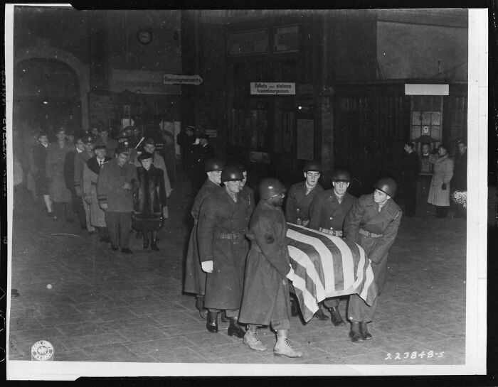 Soldiers carrying a flag-draped coffin with a mournful crowd following in a historical photo from a vastly different world.