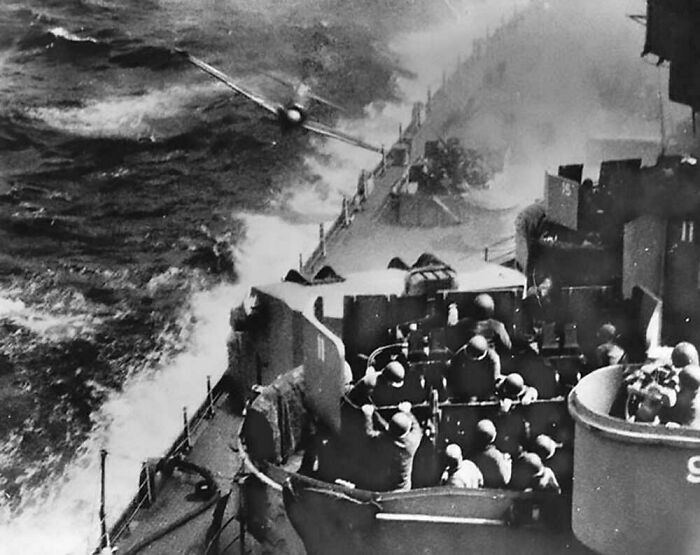 Black and white historical photo of a naval battle with soldiers on a warship and a plane flying overhead.