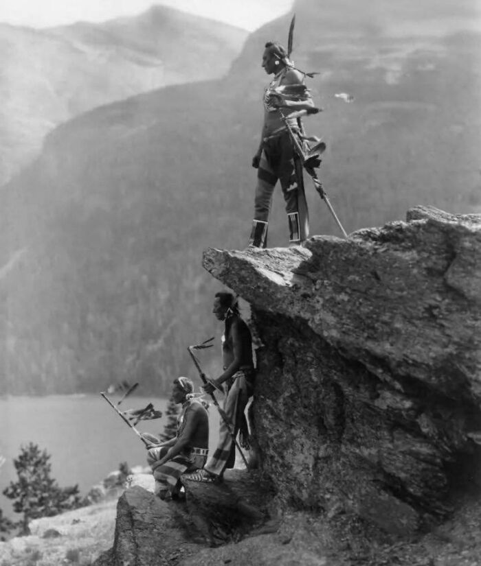 Three Native American men in traditional attire standing on a cliff captured in a historical photo representing a vastly different world.