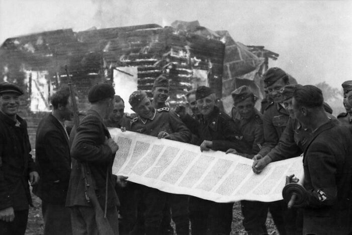 Group of soldiers and civilians examining a large document in front of a damaged wooden building in a historical photo.