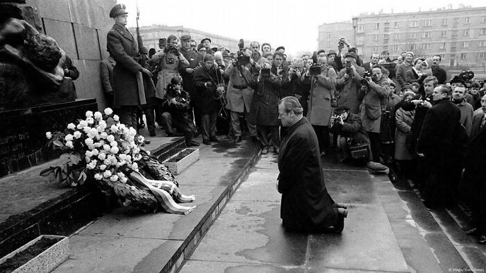 Black and white historical photo of a man kneeling at a memorial surrounded by photographers and a crowd.