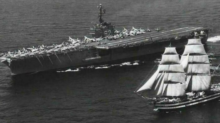Black and white historical photo showing a vintage aircraft carrier and a tall sailing ship side by side at sea.