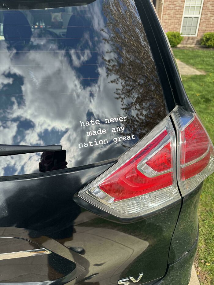 Black car window with a bumper sticker reading hate never made any nation great under a partly cloudy sky reflection.