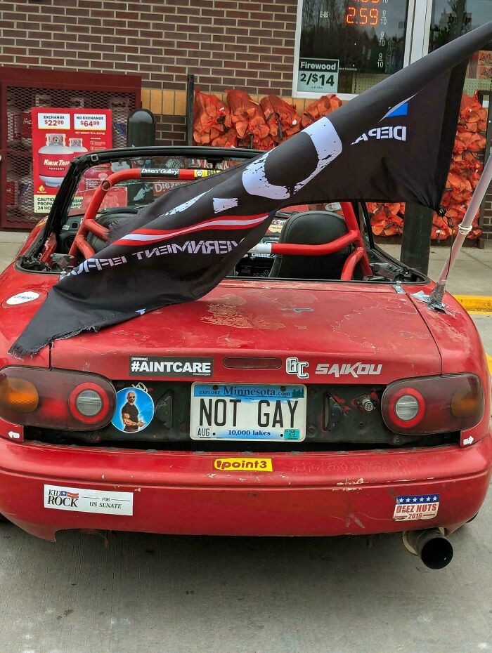 Red convertible car with humorous bumper stickers and a flag, featuring a Minnesota plate reading NOT GAY, spotted with hilarious bumper stickers.
