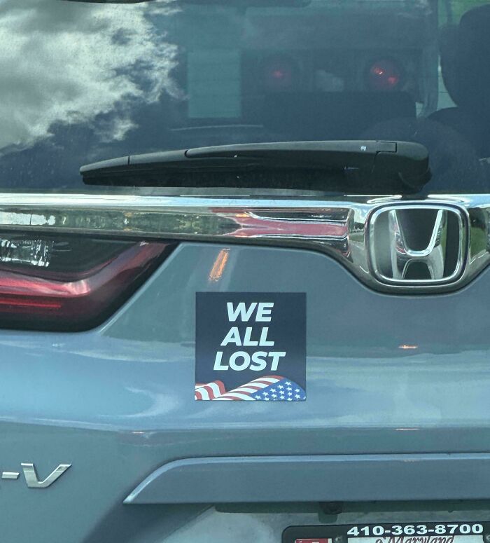 Hilarious and unhinged bumper sticker on a gray Honda SUV reading WE ALL LOST with an American flag design.