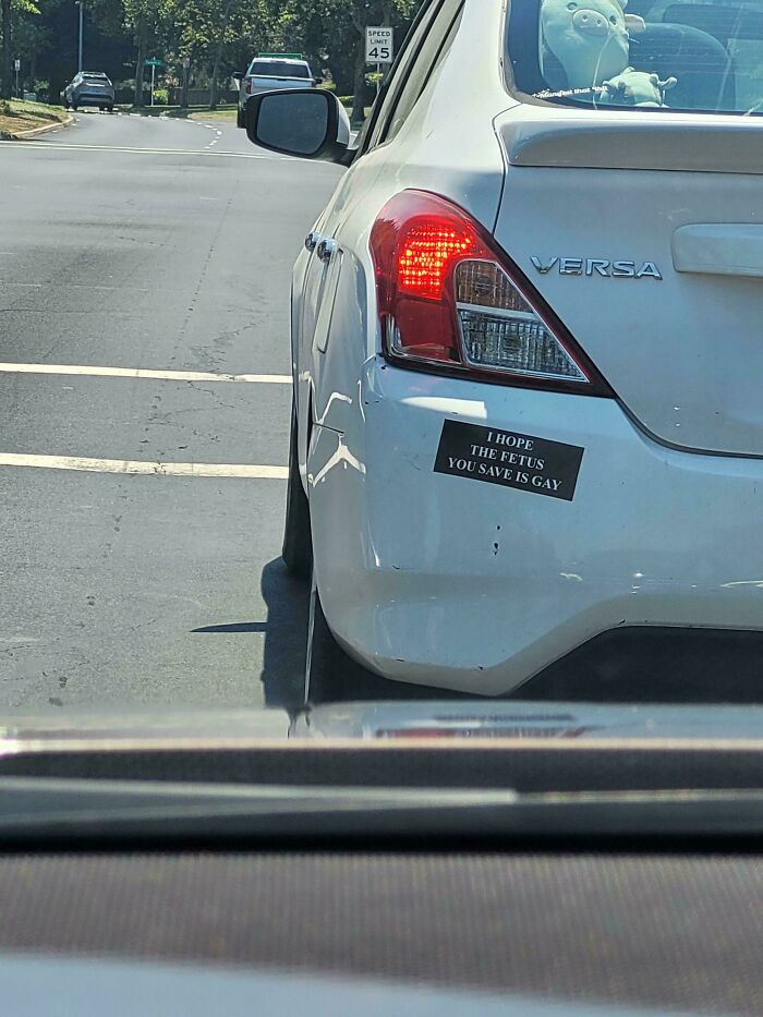 White Nissan Versa with a hilarious and unhinged bumper sticker about saving fetuses, captured on a city street.