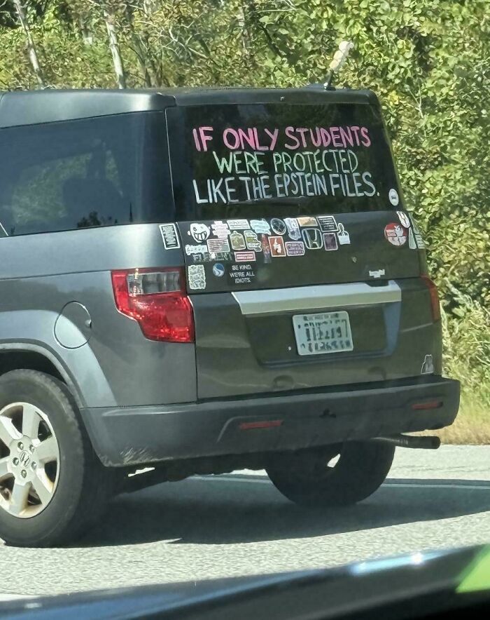 Gray SUV with multiple humorous and unhinged bumper stickers and a large text decal on rear window about students and Epstein files.