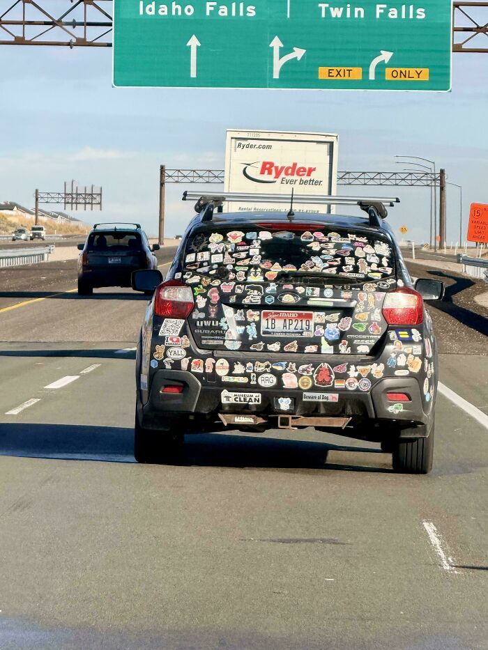 Car covered with hilarious and unhinged bumper stickers driving on highway near Idaho Falls and Twin Falls exit signs.