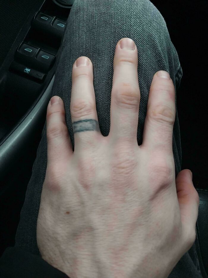 Close-up of a hand resting on gray pants showing a poorly done, blurry black ring tattoo on the finger, awful tattoos.