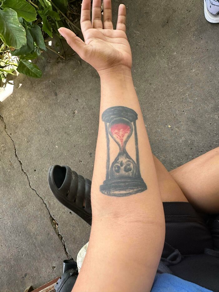 Forearm tattoo of an hourglass with a face in the sand, showcasing a truly awful tattoo with funny and tragic elements.