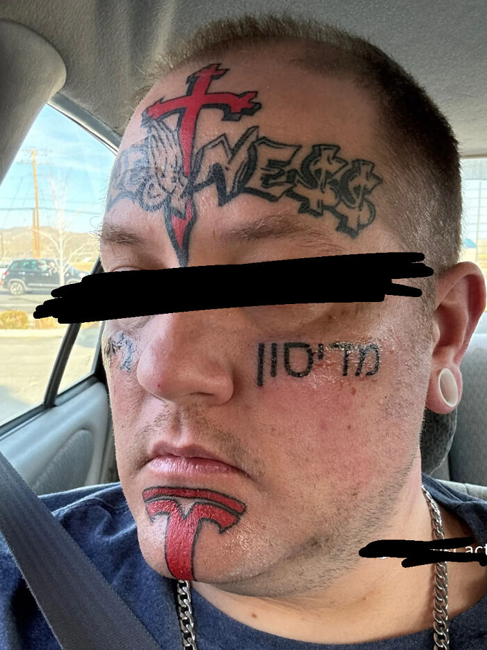 Close-up of a man with multiple truly awful tattoos on his face, including a red cross and stylized text designs.