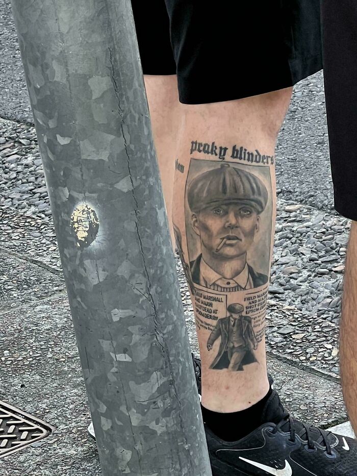Tattoo of Peaky Blinders character on calf with newspaper-style design and text in black and gray ink on a person's leg.