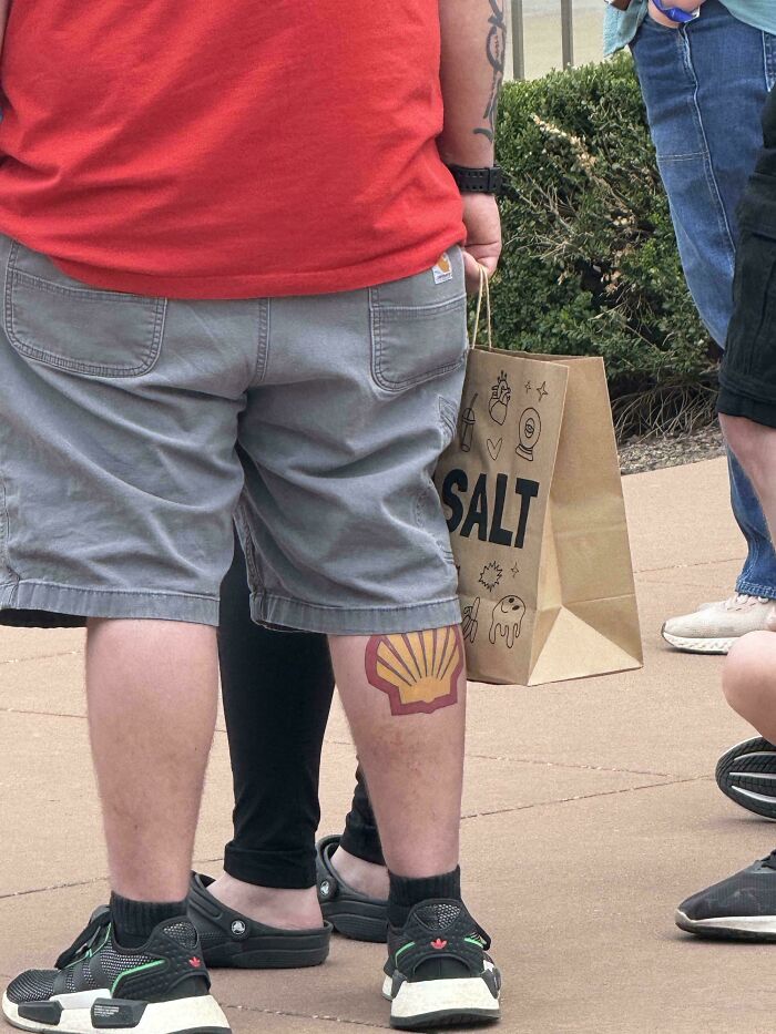 Man with large funny tattoo of a gas station logo on calf, wearing gray shorts and black sneakers outdoors with others nearby.